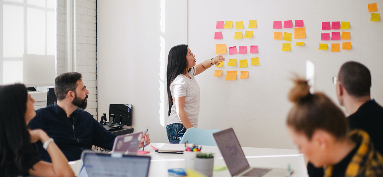 person pointing to sticky notes on wall with group of people sitting around table